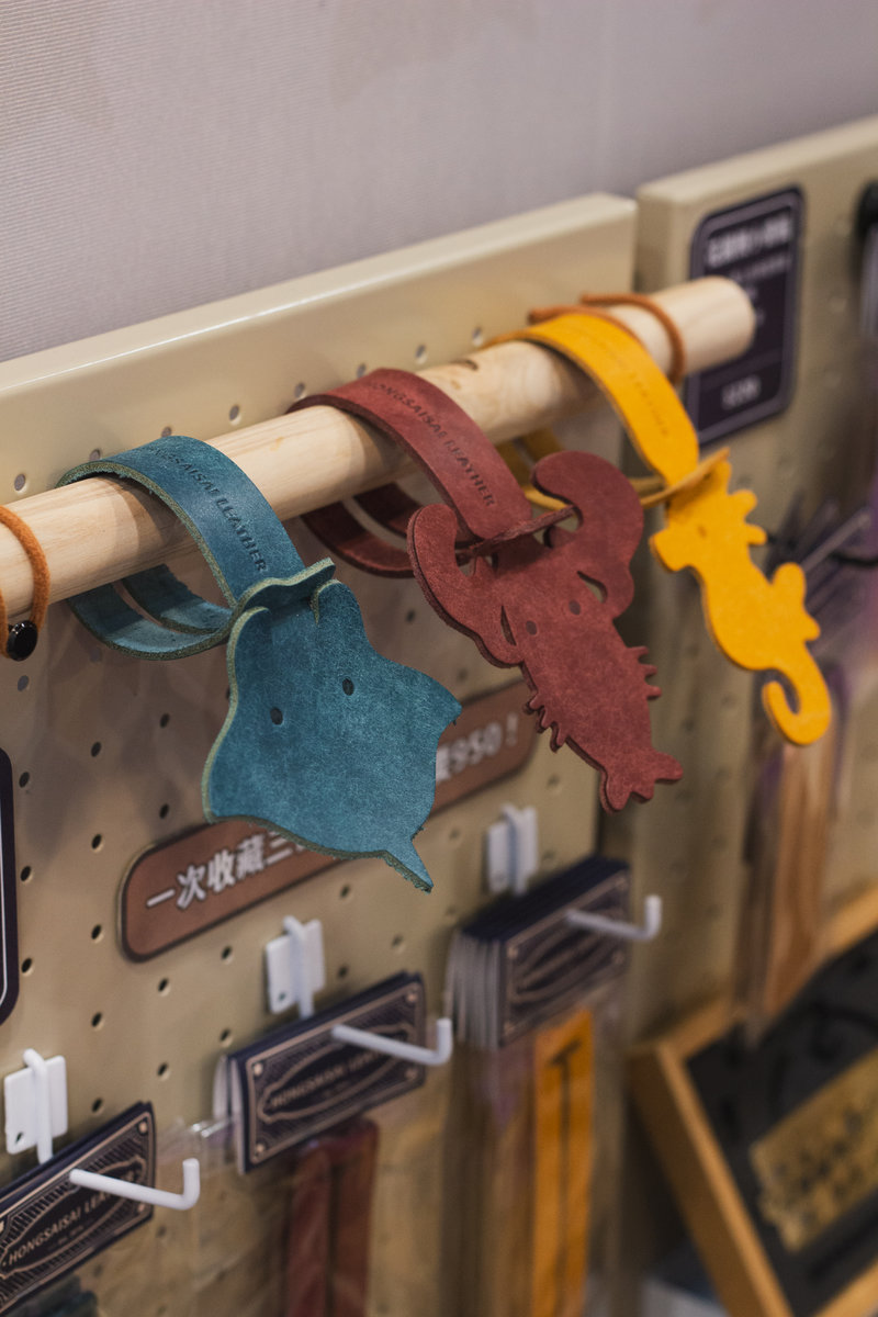 藍綠色魟魚、深紅色螃蟹、黃色海馬三款植鞣皮革行李吊牌,掛於展覽展示架木棍上,市集陳列實境場景