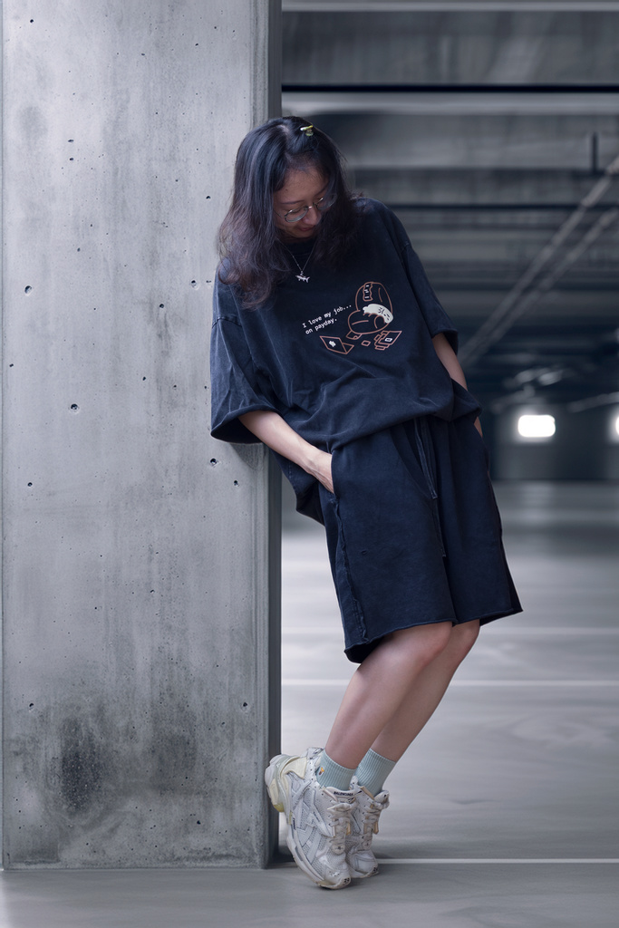 女性穿著黑色重磅T恤靠在混凝土柱上,T恤印有魟魚刺繡設計,搭配白色運動鞋於停車場拍攝