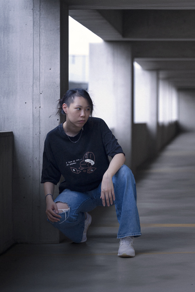女性穿著魟骰篩黑色重磅T恤搭配藍色牛仔褲,蹲坐在混凝土走廊靠牆處的穿搭展示
